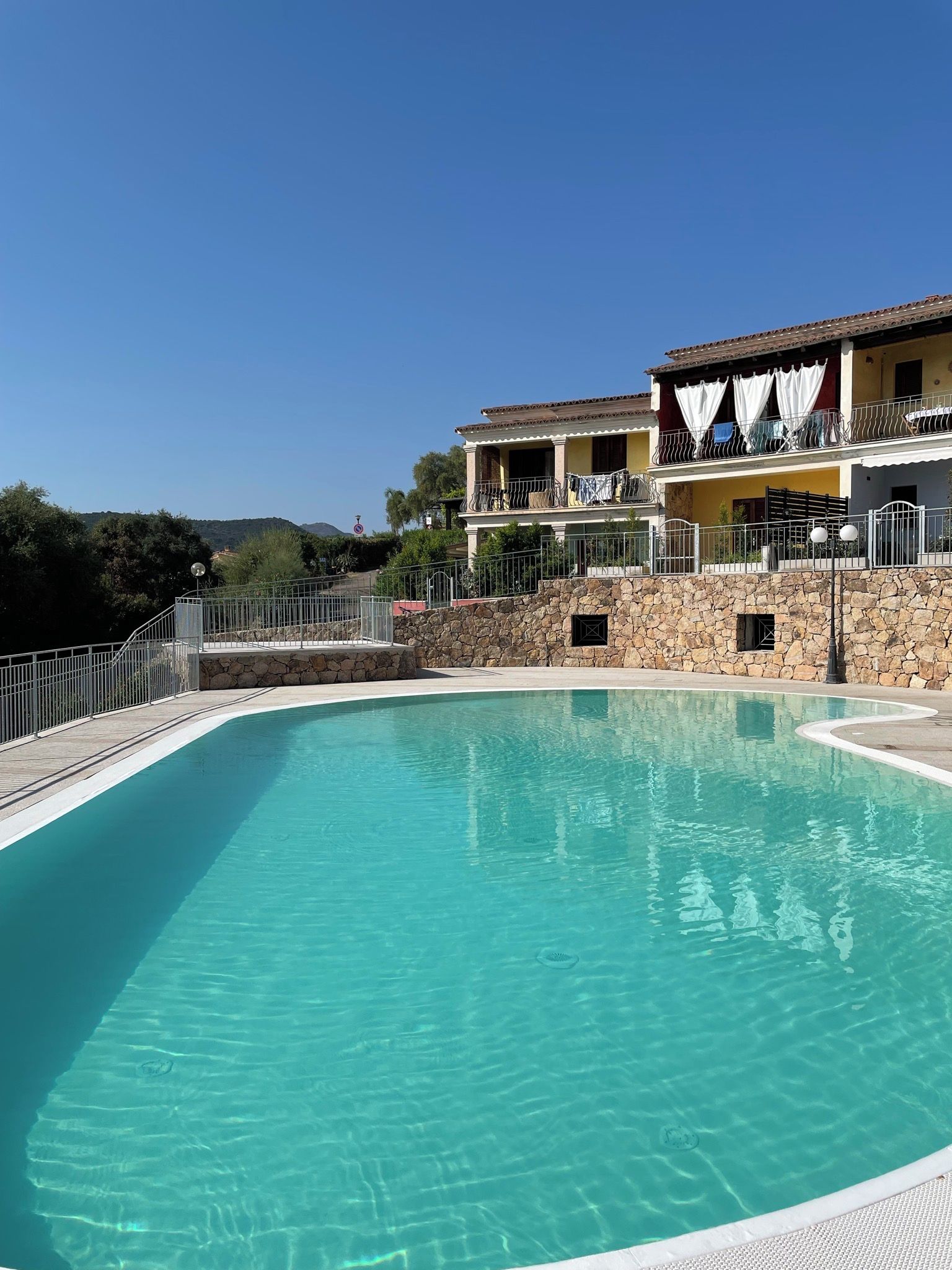 A clear swimming pool with a stone wall and a building with balconies in the background.