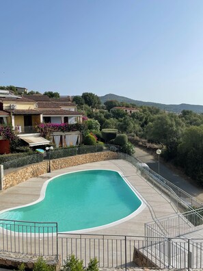 Una piscina con plataforma de piedra, rodeada por una valla metálica, y vistas a una zona residencial con casas y árboles.