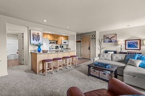 A modern kitchen with a central island, a dining area with bar stools, a living room with a sofa and coffee table, and a hallway leading to a bathroom. - Seventh Mountain Hideaway | Unit 1437 (Bend)