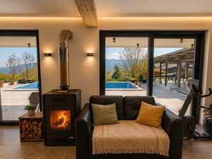 A modern living room with a wood-burning stove, a leather sofa, and a view of a swimming pool and mountains. - Luxury Hillside Villa with Pool, Terraces & Stunning Views (Gorno Sonje)