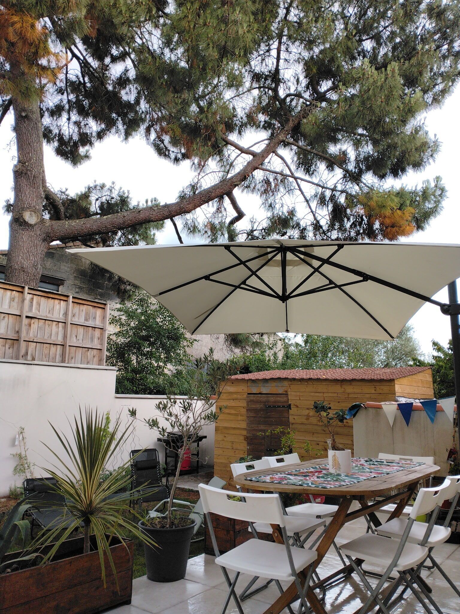 An outdoor patio with a round table, white chairs, a large umbrella, and potted plants.