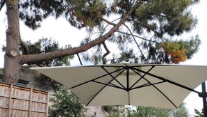 Un patio al aire libre con una mesa redonda, sillas blancas, un paraguas grande y plantas en macetas.