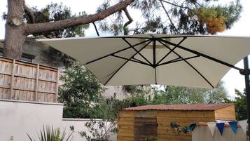 An outdoor patio with a round table, white chairs, a large umbrella, and potted plants.