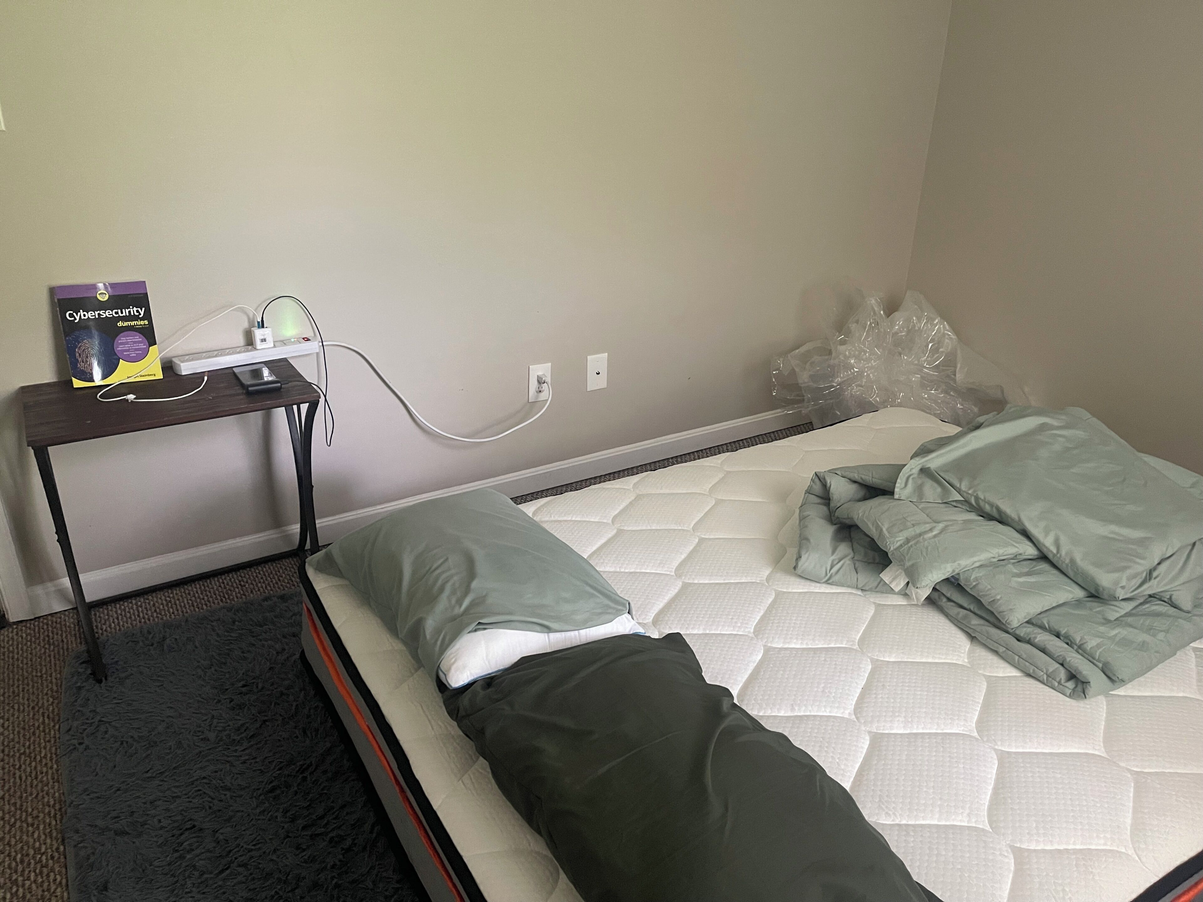 A single bed with a mattress, pillows, and a blanket. There is a small table with a book and a power strip.