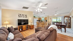 A living room with a sofa, TV, and dining area with chairs and a table. - Twisted Palms - 3/2 Dog Friendly w/ Golf Cart in The Village of Amelia (Amelia, The Villages)