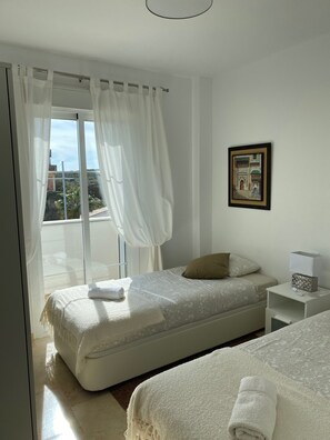 Ein Schlafzimmer mit einem Bett, einem Nachttisch, einem Fenster mit Vorhängen und einem gerahmten Bild an der Wand.