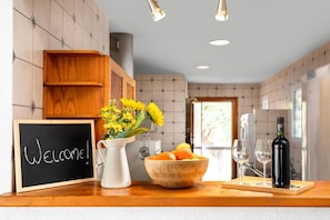 A kitchen counter with a welcome sign, a vase of flowers, a bowl of fruit, wine glasses, and a bottle of wine.