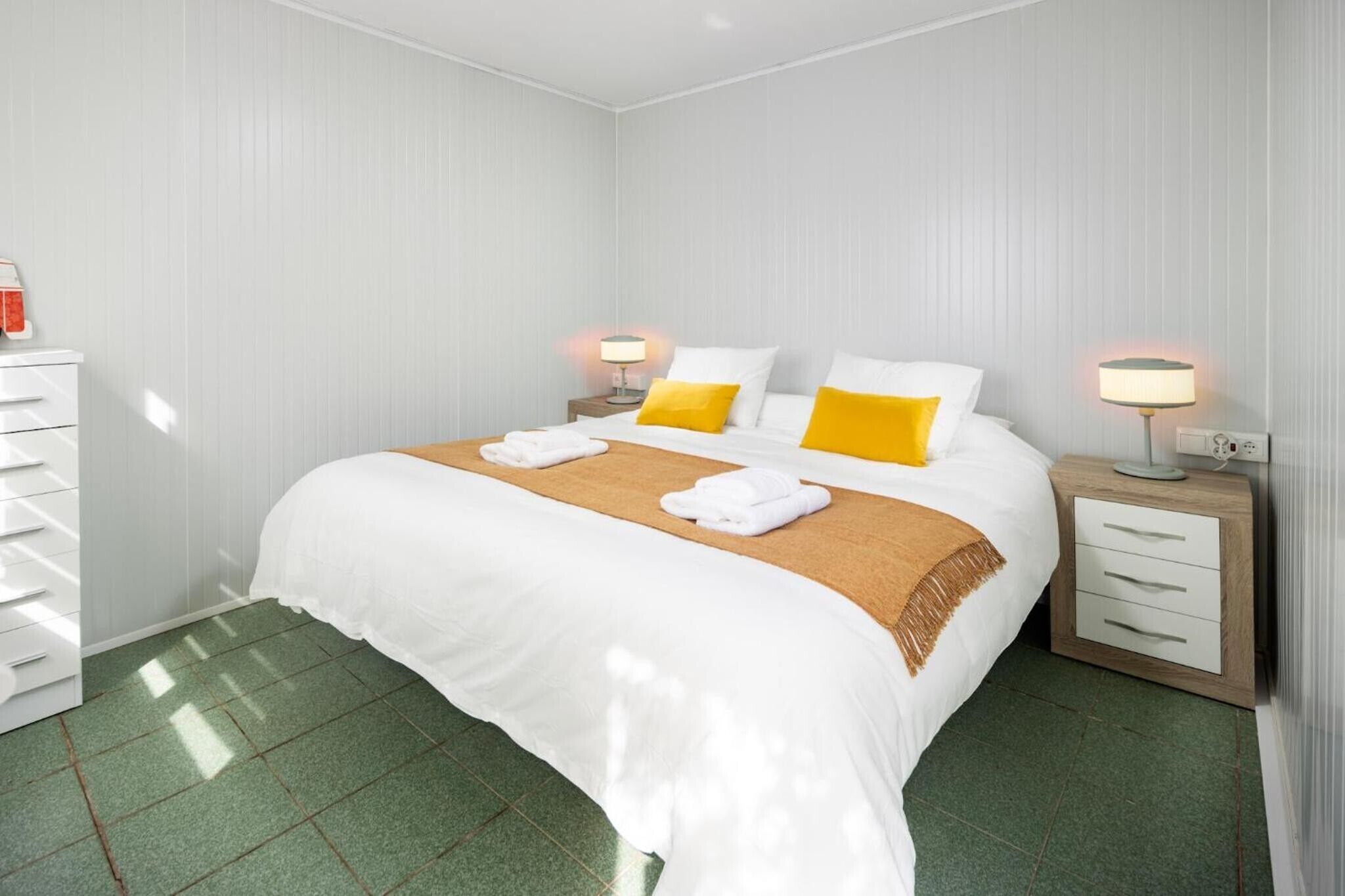Una habitación de hotel con cama, dos mesitas de noche, una cómoda y suelo de baldosas verdes.