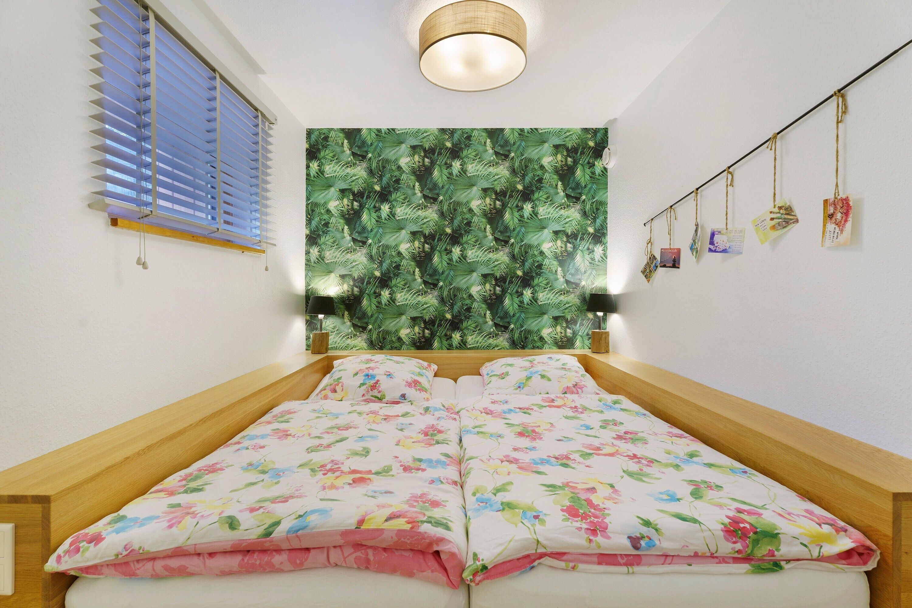 A bedroom with a bed, a window with blinds, a wall tapestry, and a string of cards.