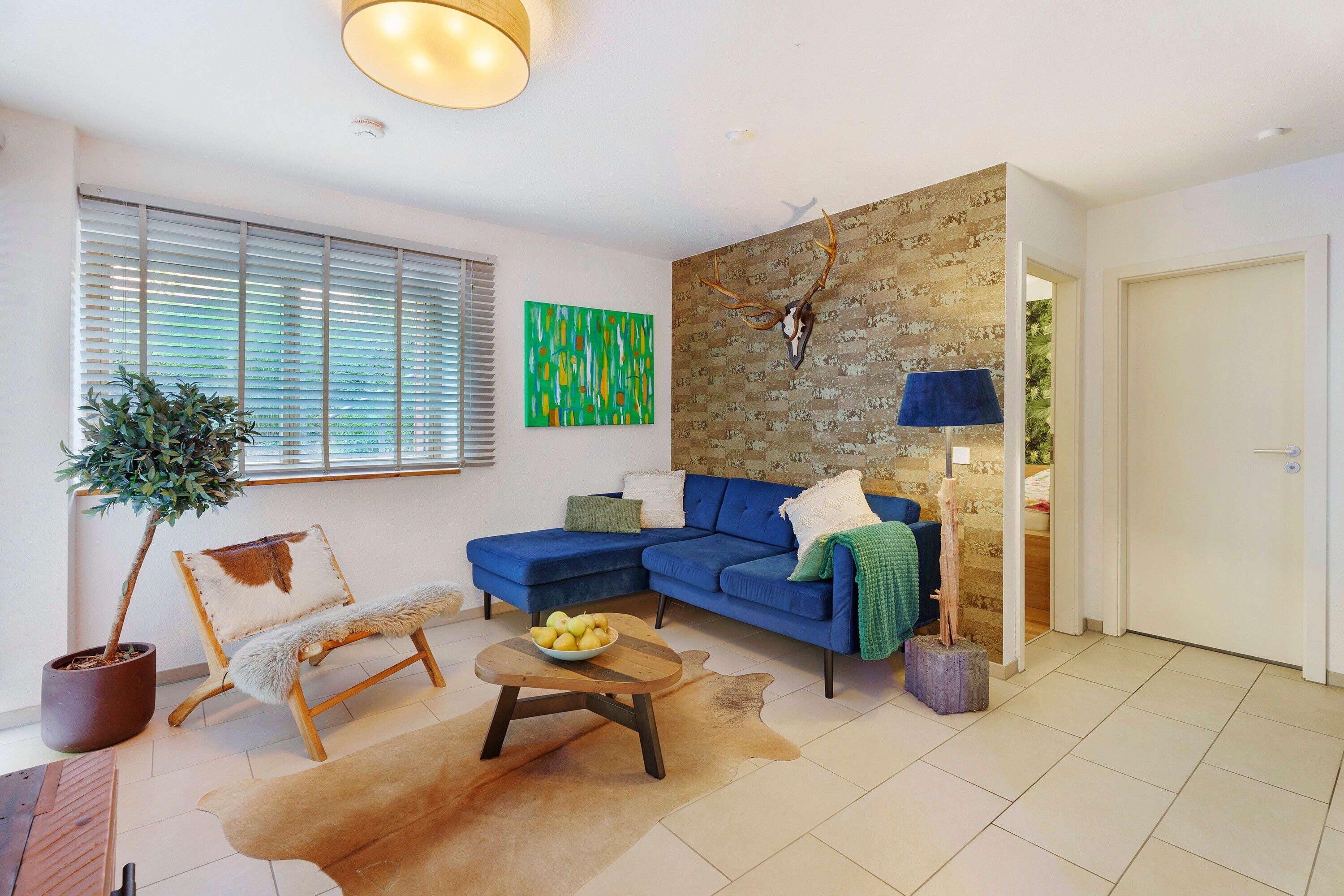 Un salon avec un canapé bleu, une table basse en bois sur laquelle se trouvent des fruits, une plante en pot, un crâne de cerf accroché au mur et un tableau abstrait vert.
