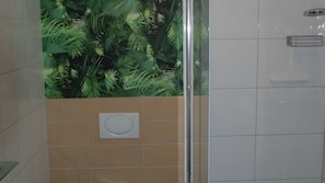 Une salle de bain avec une douche, un mur doté d’une grille de ventilation circulaire et un mur carrelé orné d’un motif de feuilles tropicales.