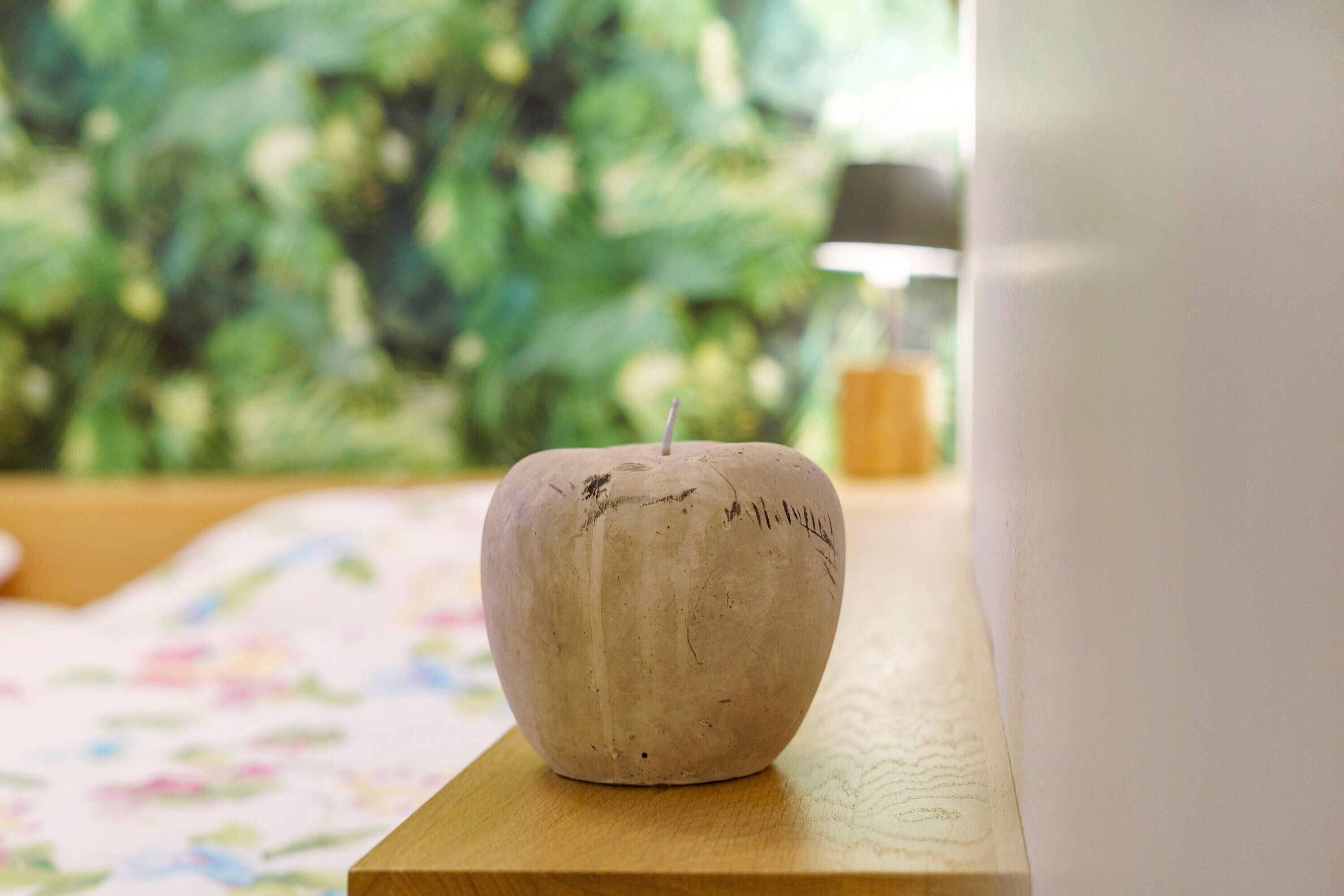 Ein Holztisch mit einem Kerzenständer in Apfelform, einer Tischdecke mit floralem Muster und einem grünen, blättrigen Hintergrund.