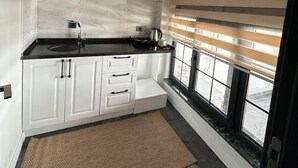A modern kitchen with white cabinets, a black countertop, a hanging light fixture, and a window with blinds. - Ay-Asia Suit (Amasya)