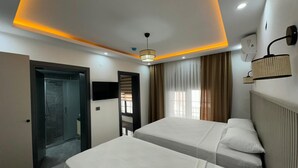 A hotel room with two beds, a TV, a ceiling light, a window with curtains, and a door leading to a bathroom. - Ay-Asia Suit (Amasya)