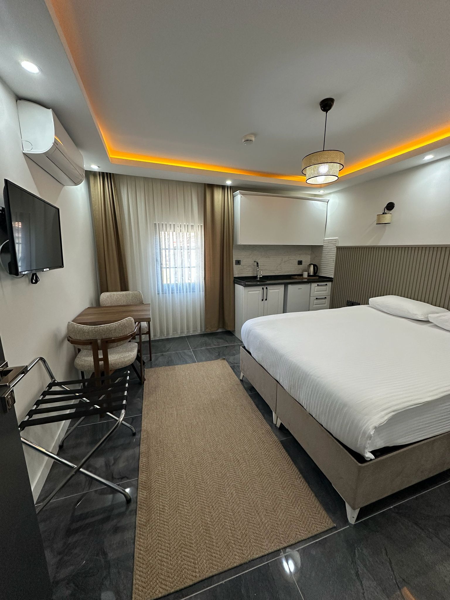 Quarto de hotel moderno com cama, escrivaninha, cadeira, televisão e cozinha compacta.