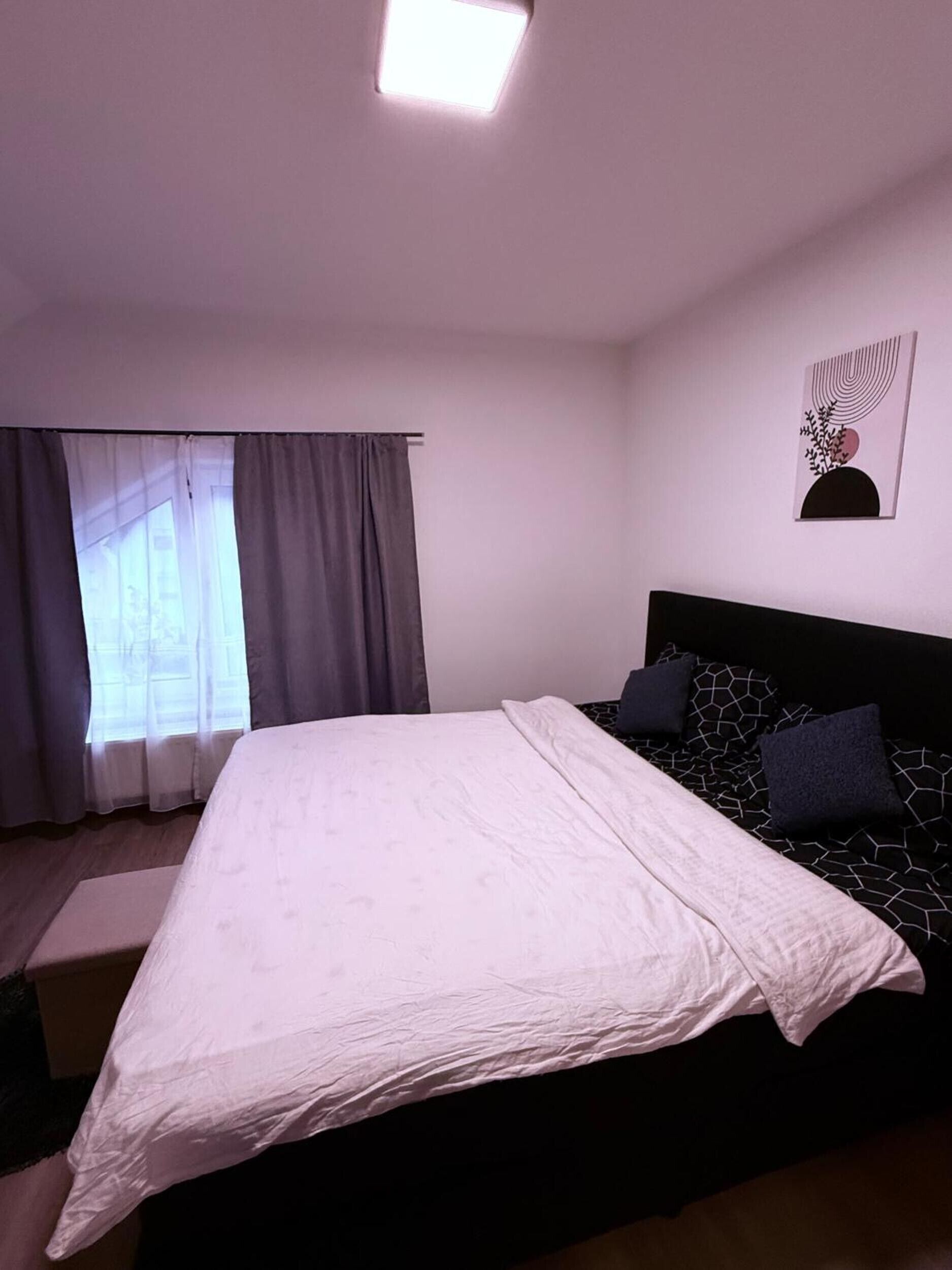 A bedroom with a bed, a window with curtains, a headboard, and a wall-mounted artwork.
