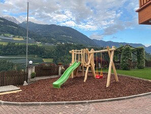 Une aire de jeux avec un toboggan, des balançoires et une structure en bois, entourée de paillis et bordée de briques, avec un paysage montagneux en arrière-plan.