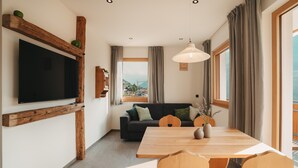 Moderni olohuone, jossa on litteänäyttöinen televisio, puupalkit, sohva sekä ruokailutila, jossa on pöytä ja tuolit.