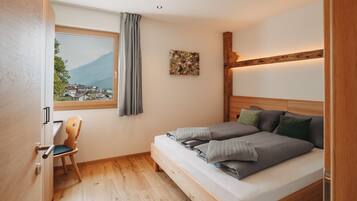 A bedroom with a bed, a chair, a window with a view of houses and mountains, and a painting on the wall.