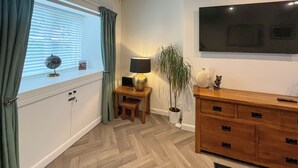 A room with a wooden herringbone floor, a TV mounted on the wall, a wooden cabinet with drawers, a small table with a lamp, a potted plant, and a globe on the windowsill.