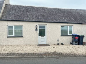 A house with a white door, two windows with shutters, a small porch, and a gravel driveway.