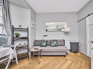 A modern living room with a grey sofa, a small round table, a white desk, a standing lamp, and a trash bin. - No.1 Princess Blue Apartments (Portrush)