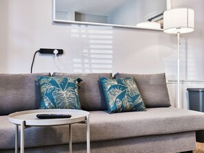 A modern living room with a grey sofa, two patterned throw pillows, a remote control on a small table, and a floor lamp. - No.1 Princess Blue Apartments (Portrush)
