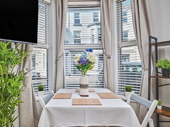 A room with a table set for dining, a vase with flowers, and a view of buildings through large windows. - No.1 Princess Blue Apartments (Portrush)