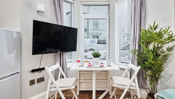 A corner room with a flat-screen TV, two white chairs, a small table with a plant, and a window with blinds. - No.1 Princess Green Apartments - UK52036 (Portrush)
