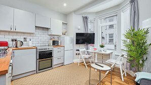 A modern kitchen with white cabinets, a built-in oven, a microwave, and a refrigerator. There is a dining area with a table and chairs, a TV mounted on the wall, and a large window with blinds. - No.1 Princess Green Apartments - UK52036 (Portrush)