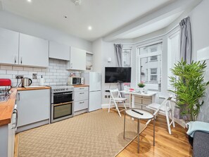 A modern kitchen with white cabinets, a built-in oven, a microwave, and a refrigerator. There is a dining area with a table and chairs, a TV mounted on the wall, and a large window with blinds. - No.1 Princess Green Apartments - UK52036 (Portrush)