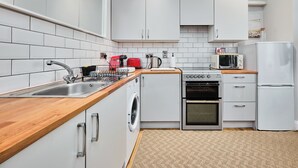 A modern kitchen with white cabinets, a stainless steel sink, a washing machine, a stove, a microwave, and a refrigerator. - No.1 Princess Green Apartments - UK52036 (Portrush)
