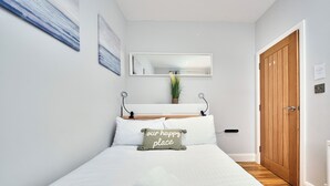 A neatly arranged bedroom with a bed, pillows, a bedside table, a mirror, a wooden door, and a small potted plant. - No.1 Princess Green Apartments - UK52036 (Portrush)
