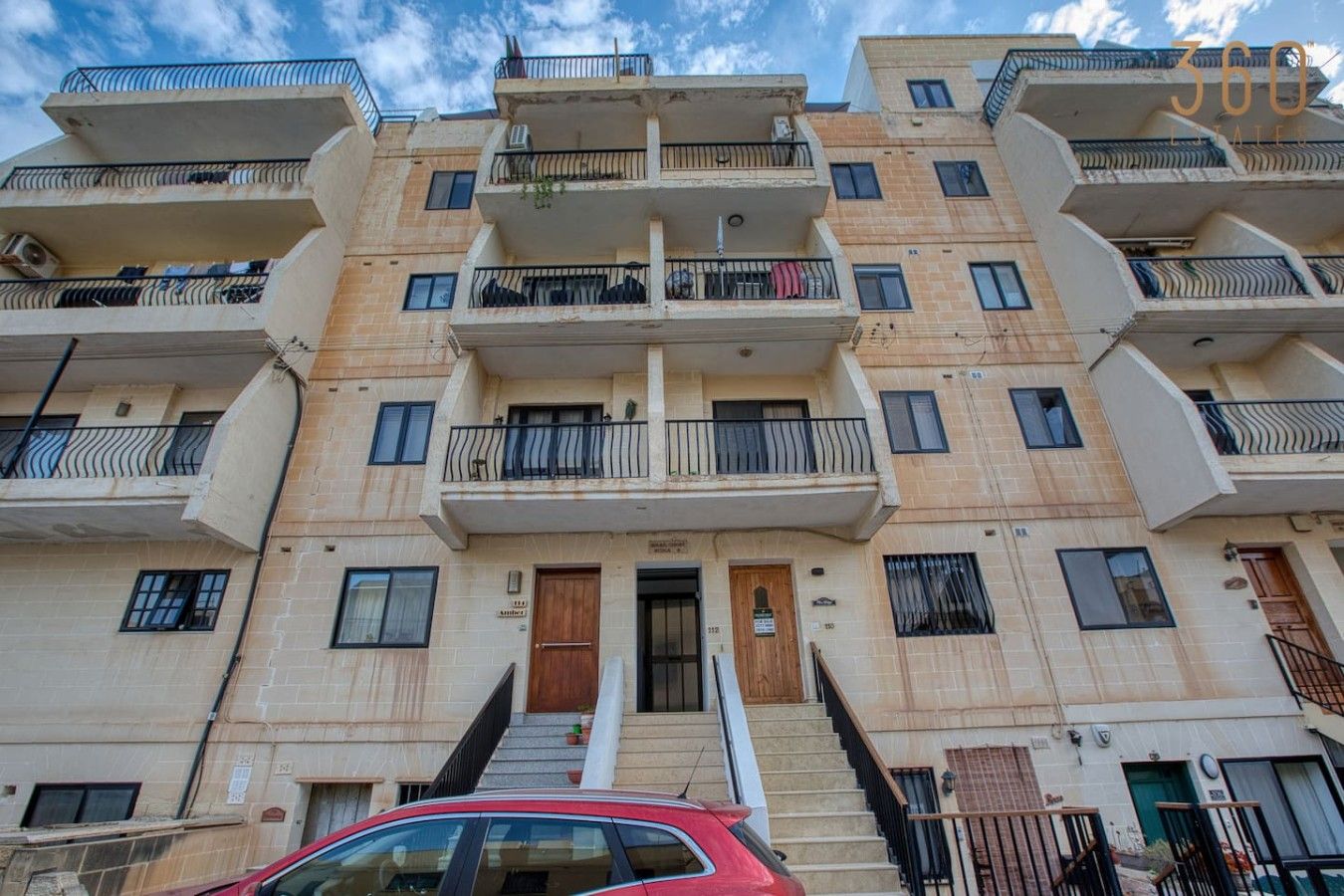 A multi-story residential building with balconies, a central staircase, and a red car parked in front.
