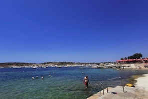 A beach with clear water, people swimming, and a row of red umbrellas. - A Premium Apartment in the Heart of Marsaskala (Marsascala MALTA)