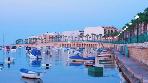 A marina with boats docked along a waterfront promenade, featuring a stone wall and palm trees in the background. - A Premium Apartment in the Heart of Marsaskala (Marsascala MALTA)