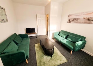 Uma sala de estar com dois sofás verdes, uma mesa de centro e uma TV sobre um móvel.