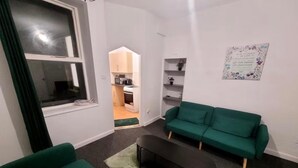 A living room with a green sofa, a black coffee table, and a kitchen visible through an open door. - Cozy Apartment in Barrow-in-furness (Barrow-in-Furness)