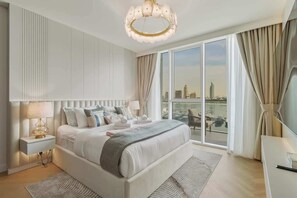 A modern bedroom with a large bed, a nightstand, a lamp, a view of the city through large windows, and a chandelier. - Sea-View 1BR + Private Beach, Beach Mansion (Dubai)