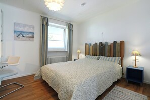 A bedroom with a bed, bedside tables, a chair, a window with curtains, a painting on the wall, and a lamp. - House Wendt Apartment 01 (Timmendorfer Strand)