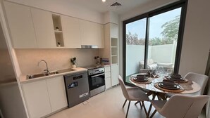 A modern kitchen with built-in appliances, a dining area with a table set for four, and a large window providing a view of the outdoors. - Sun-Drenched 3BR Garden Villa, Arabian Ranches (Dubai)