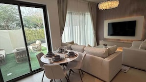 A modern living room with a sofa set, a dining table with chairs, a TV mounted on the wall, and a large sliding glass door leading to an outdoor area. - Sun-Drenched 3BR Garden Villa, Arabian Ranches (Dubai)