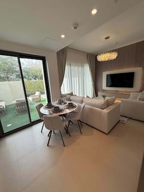 Ein modernes Wohnzimmer mit einer Sofagarnitur, einem Esstisch mit Stühlen, einem an der Wand montierten Fernseher und einer großen Glasschiebetür, die zu einem Außenbereich führt.
