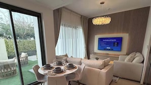 A modern living room with a dining area, a flat-screen TV mounted on the wall, and a view of the outdoors. - Sun-Drenched 3BR Garden Villa, Arabian Ranches (Dubai)