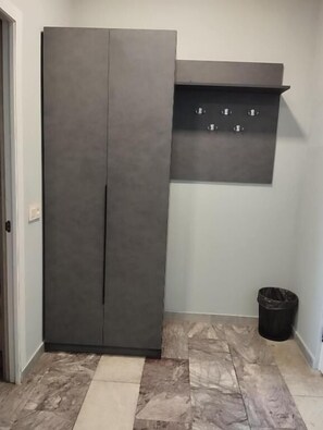 Quarto com um guarda-roupa cinzento, uma pequena prateleira e um cesto de lixo.
