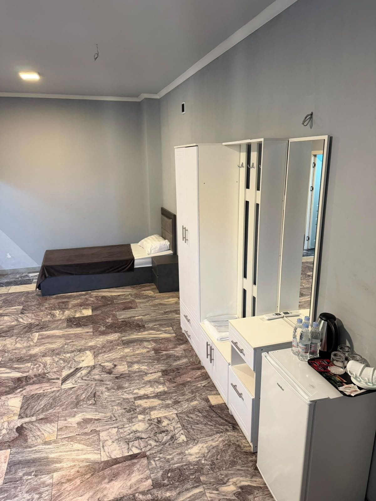 A bedroom with a bed, wardrobe, mirror, and a small kitchenette area.