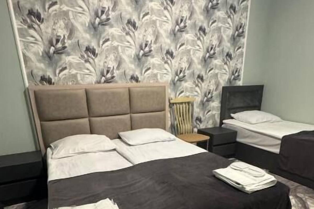 Una habitación de hotel con una cama grande, una cama más pequeña, una mesita de noche, una lámpara y un papel tapiz con motivos florales.