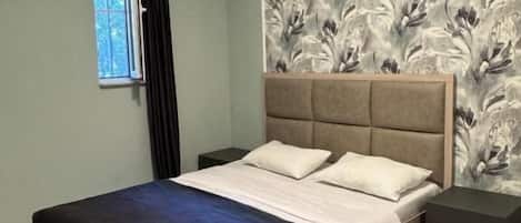 A bedroom with a bed, a headboard, a nightstand, a window, and a wall with a floral pattern.
