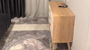A room with a TV mounted on the wall, a wooden dresser, a window with sheer curtains, and a marble-patterned floor. - Corner Hotel  (Yerevan)
