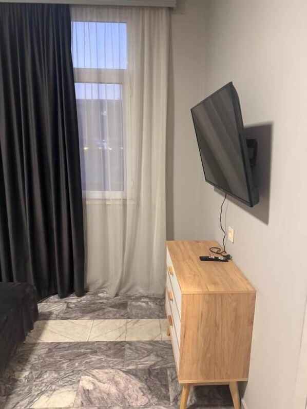 Ein Zimmer mit an der Wand montiertem Fernseher, einem hölzernen Schminktisch, einem Fenster mit durchsichtigen Vorhängen und einem Boden mit Marmoroptik.